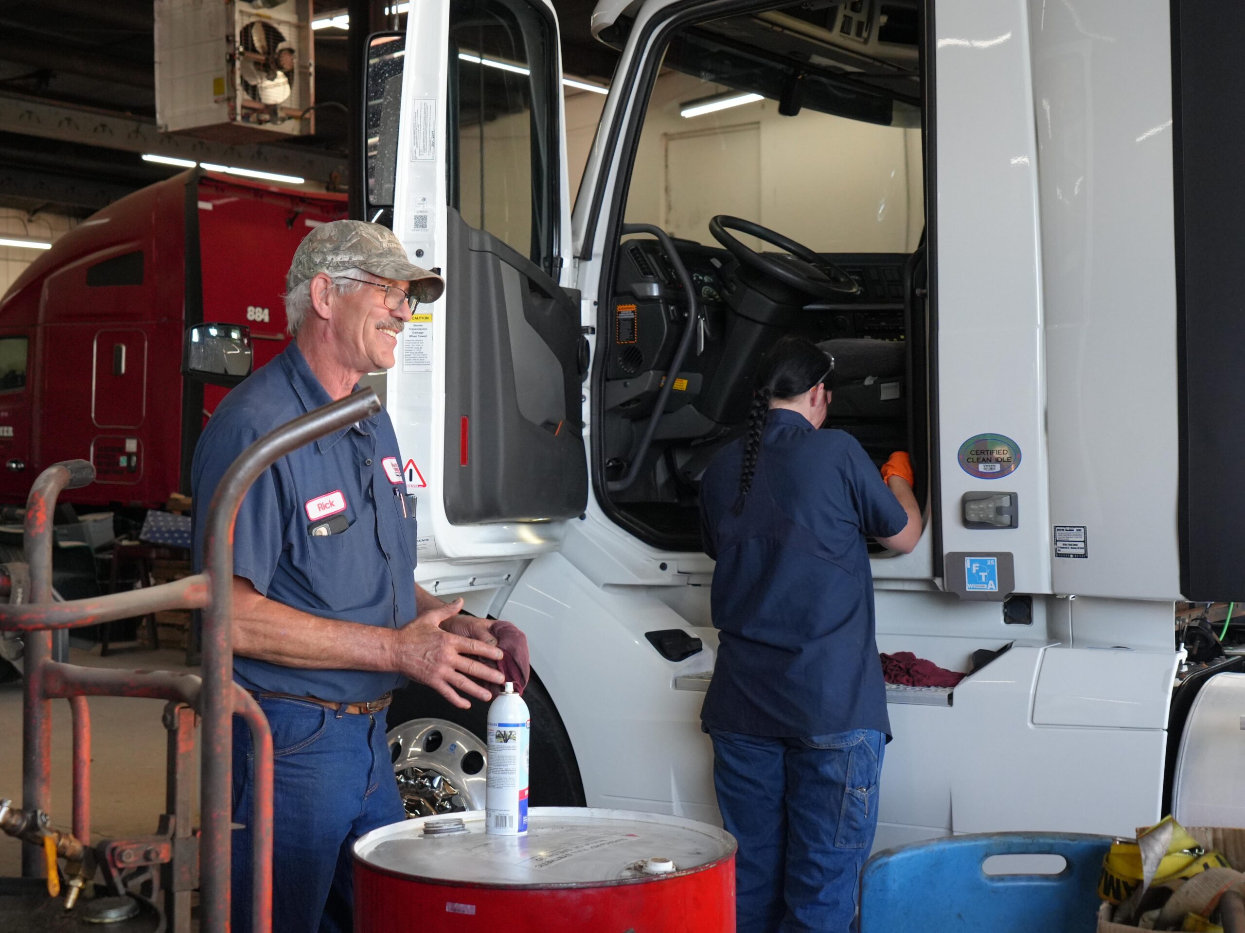 A truck mechanic stands beside a white semi-truck, smiling while a colleague works inside the cab, both engaged in maintenance tasks.