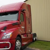 Red semi-truck parked close to a yellow warehouse, showcasing its sleek design and powerful build. Relevant for discussions on transportation or logistics.