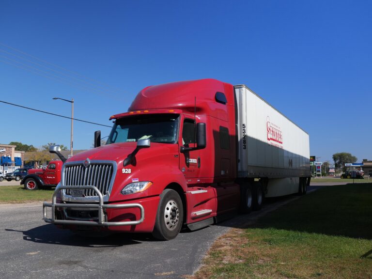 Red semi-truck with a white trailer parked on a road, surrounded by greenery and nearby businesses, showcasing transportation logistics.