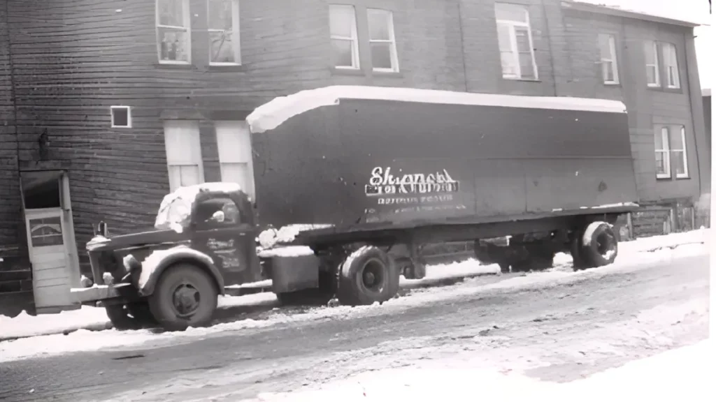 Vintage truck covered in snow, parked on a street beside a wooden building. The truck advertises a local delivery service, reflecting mid-20th century transport.