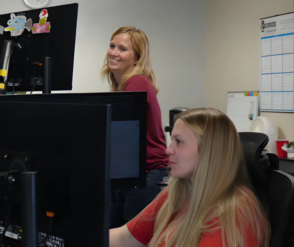 Two women work at computers in an office setting, smiling and engaged. One stands behind the other, fostering a collaborative atmosphere.