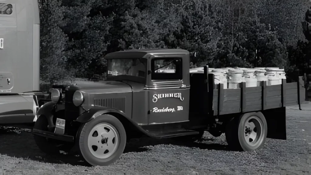 Classic black-and-white photo of a vintage truck with a wooden bed, loaded with jars, parked amidst greenery. Represents retro transportation and local craftsmanship.