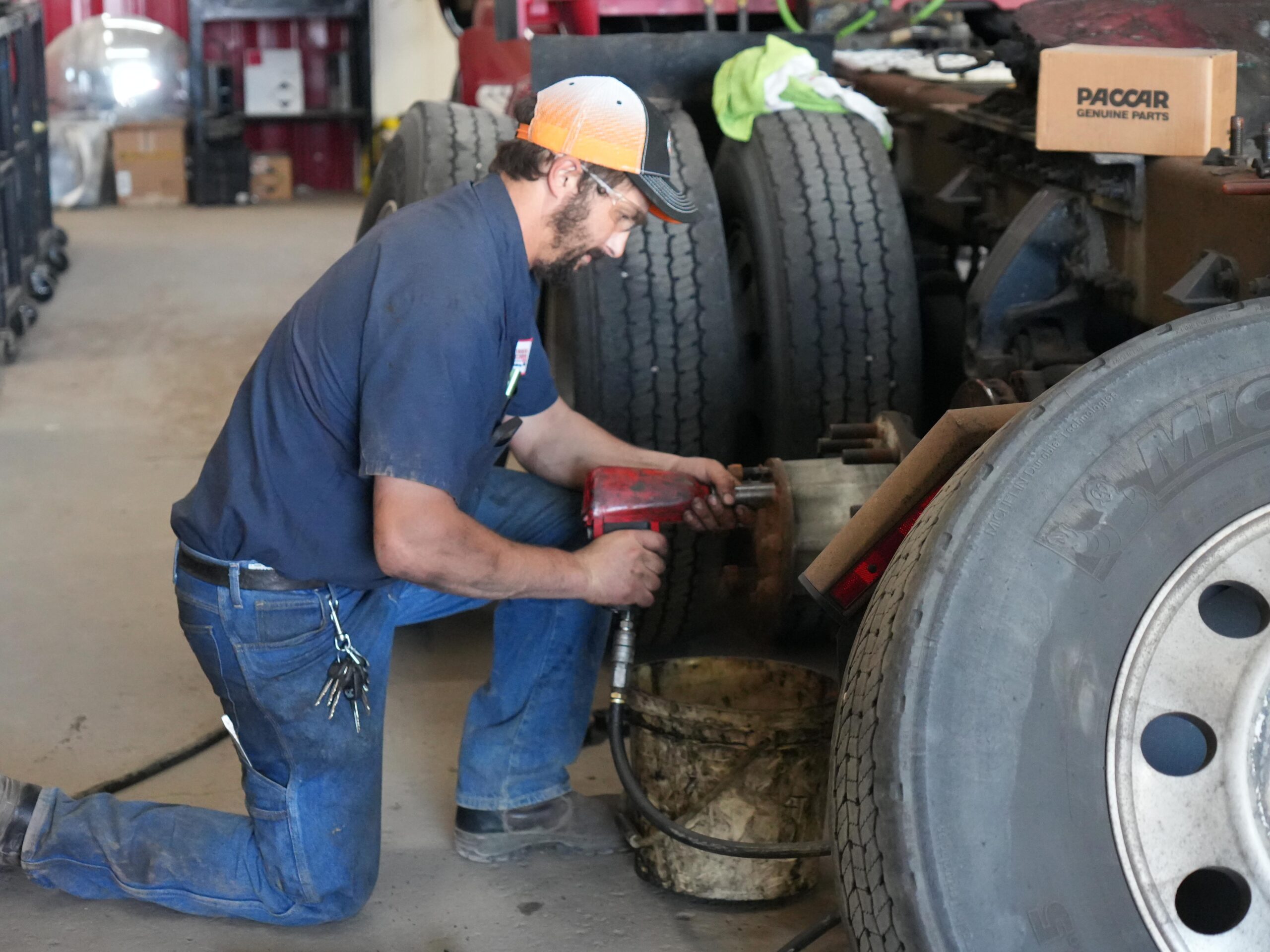 A mechanic kneels beside a large truck in a repair shop, using an air impact wrench to work on the wheel assembly. Tools and parts are visible in the background.